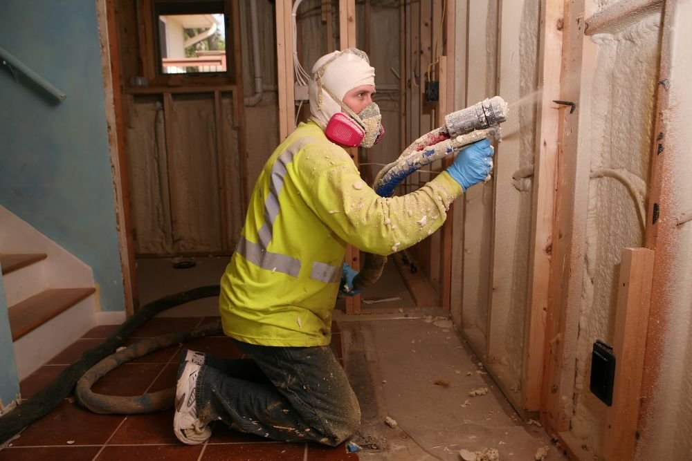 A construction worker in a yellow safety vest and mask kneels while applying insulation between wooden frames in a home renovation.