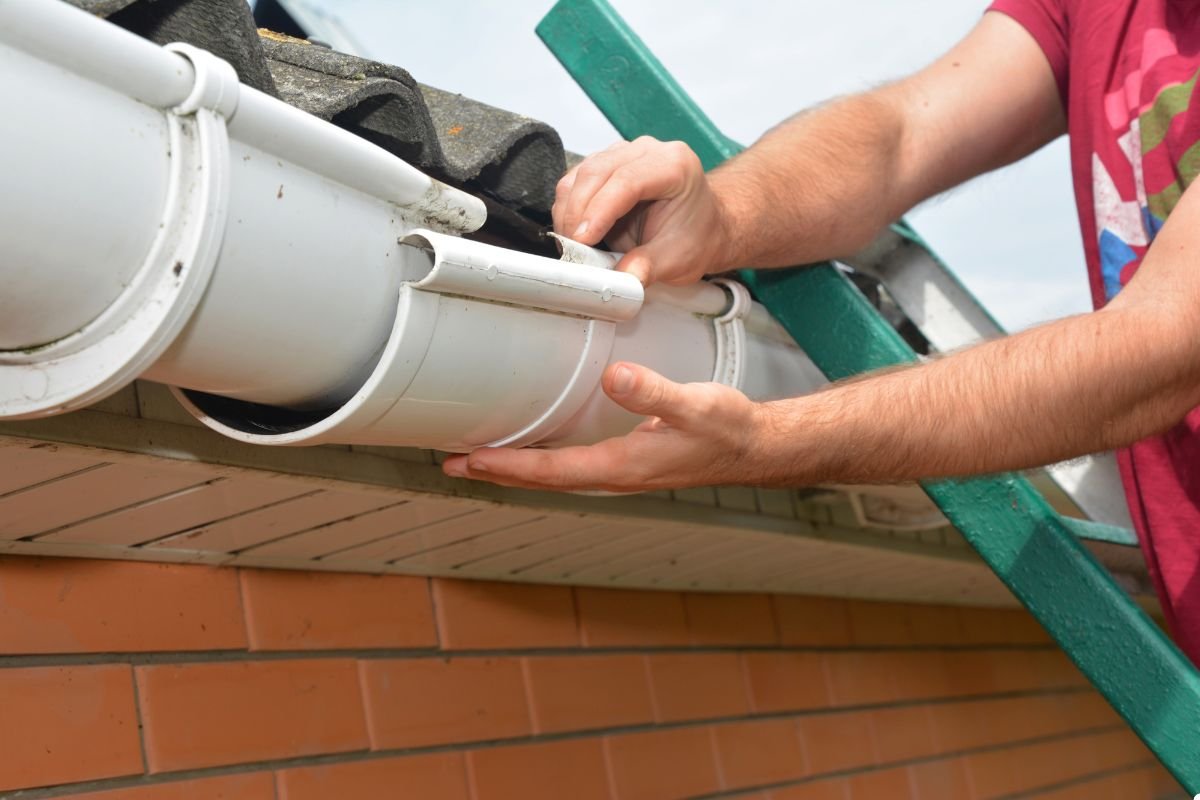 A man is on a ladder, fixing a gutter on a brick wall, focused on ensuring proper drainage and maintenance.