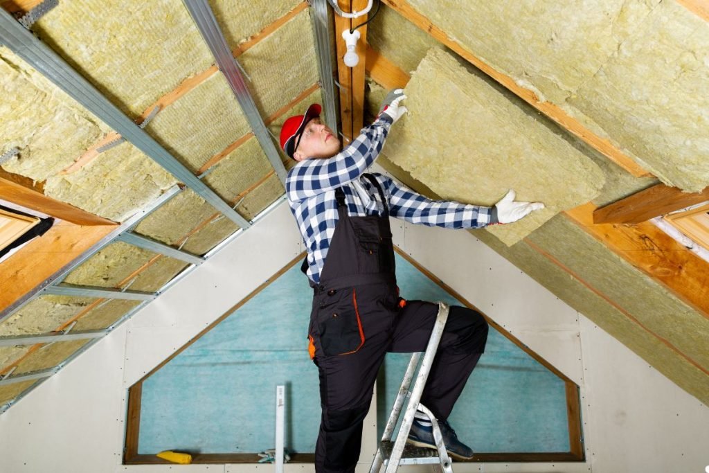 A man installs insulation on a roof, wearing safety gear and focused on his task under a clear blue sky.