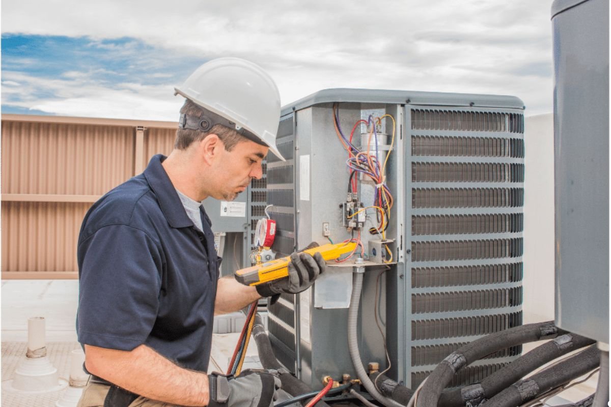 A man in a blue shirt repairs an air conditioner, focused on the unit with tools in hand.
